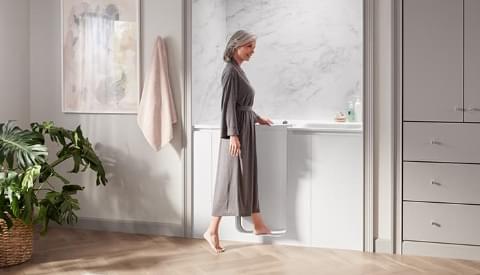 far away view of woman in gray robe entering her bath stepping over the low step entry
